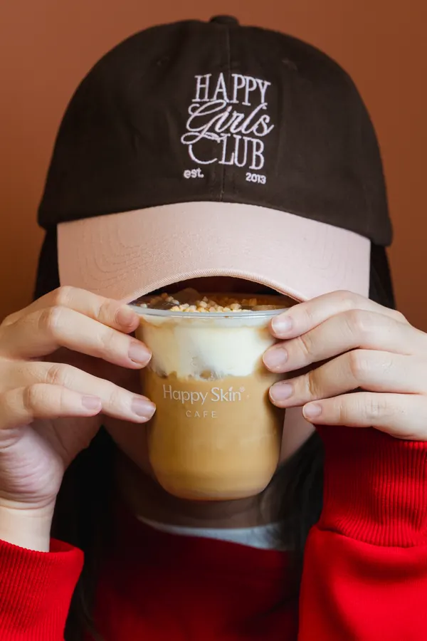Drink held up in front of a Happy Girls Club cap